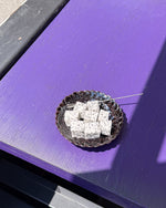 A small dark bowl containing several white speckled cubed fruits, placed on a bright purple table outdoors with strong sunlight and shadows.