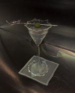A clear martini glass filled with a pale drink, garnished with a green olive on a thin metal skewer, resting on a square gray napkin on a dark reflective surface.