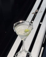 A clear martini glass filled with a pale drink and garnished with a green olive and a decorative metal pick, set against a dark and light slatted background.