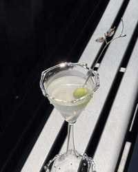 A clear martini glass filled with a pale drink and garnished with a green olive and a decorative metal pick, set against a dark and light slatted background.