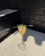 A clear stemmed glass filled with a pale yellow cocktail, garnished with a green olive on a metal pick, set on a concrete surface against a dark brick wall in bright sunlight.