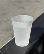 A translucent frosted glass cup with condensation droplets, sitting on a dark metal ledge outdoors against a gray concrete background.