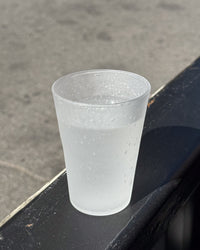 A translucent frosted glass cup with condensation droplets, sitting on a dark metal ledge outdoors against a gray concrete background.
