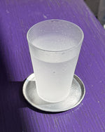 A frosted glass tumbler with condensation droplets, filled with a clear liquid, resting on a round silver coaster on a purple wooden surface.