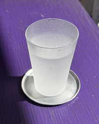 A frosted glass tumbler with condensation droplets, filled with a clear liquid, resting on a round silver coaster on a purple wooden surface.
