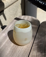 A frosted glass tumbler with small raised bubble details, filled with a pale yellow iced drink and ice cubes, resting on a weathered gray wooden table in outdoor sunlight.