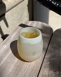 A frosted glass tumbler with small raised bubble details, filled with a pale yellow iced drink and ice cubes, resting on a weathered gray wooden table in outdoor sunlight.