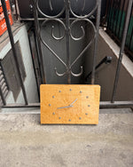 A minimalist wooden wall clock with a natural light wood face and silver metal hands, leaning against a black wrought-iron gate on a concrete surface.