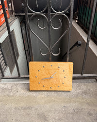 A minimalist wooden wall clock with a natural light wood face and silver metal hands, leaning against a black wrought-iron gate on a concrete surface.