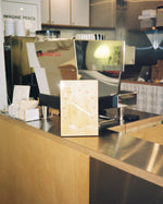 A rectangular wall clock with a light wood grain face and silver hands, styled on a stainless steel cafe counter in front of espresso machines.