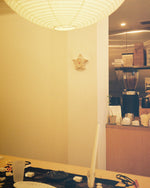 A warm, softly lit interior corner with a large white paper lantern hanging from the ceiling, a small star-shaped wall decor piece, and a counter area with a coffee maker and cups in the background.