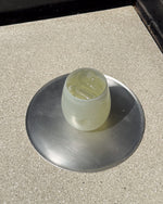 A glass tumbler containing a pale yellow liquid and ice, sitting on a round brushed metal coaster over a speckled beige countertop.