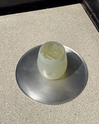 A glass tumbler containing a pale yellow liquid and ice, sitting on a round brushed metal coaster over a speckled beige countertop.