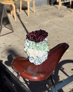 A stack of four satin hair scrunchies in burgundy, mint green, cream, and light blue is styled on top of a brown bicycle saddle outdoors on a concrete surface.