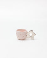 A small pastel pink ceramic espresso or tea cup with a star-shaped handle, featuring subtle embossed star patterns, set against a clean white background.