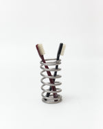 A silver metal spiral toothbrush holder containing two toothbrushes, one with a dark red handle and one with a black handle, set against a plain white background.