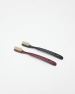 Two small toothbrushes on a clean white background, one with a dark burgundy handle and one with a black handle, both with cream bristles. The handles are printed with the brand name 'ACCA KAPPA'