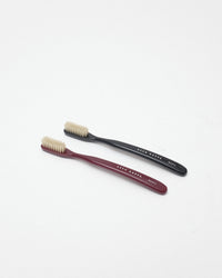Two small toothbrushes on a clean white background, one with a dark burgundy handle and one with a black handle, both with cream bristles. The handles are printed with the brand name 'ACCA KAPPA'