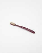 A single toothbrush with a dark burgundy handle and cream-colored bristles, featuring white printed text on the handle, photographed on a seamless white background.