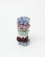 A stack of seven satin hair scrunchies in muted pastel and earthy colors, including dusty blue, pink, sage green, melon green, cream, burgundy, and dusty blue set against a plain white background.