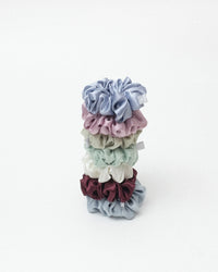 A stack of seven satin hair scrunchies in muted pastel and earthy colors, including dusty blue, pink, sage green, melon green, cream, burgundy, and dusty blue set against a plain white background.