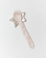 A light pink ceramic spoon with a star-shaped handle, photographed on a plain light gray background.