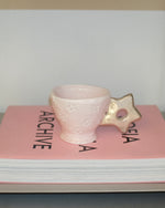 A pale pink ceramic cup with embossed star details and a star-shaped handle, resting on top of a pink book.