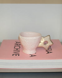 A pale pink ceramic cup with embossed star details and a star-shaped handle, resting on top of a pink book.