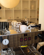 A stainless steel espresso machine with a glossy reflective surface, featuring a pink ceramic handle-shaped mug resting on its upper platform, alongside other mugs and glasses.