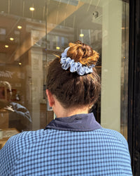 A person viewed from behind wearing a light blue scrunchie in their brown hair, standing in front of a glass storefront.