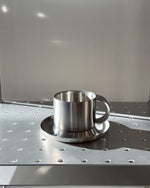 Stainless steel cup and saucer on a metallic surface with a neutral background