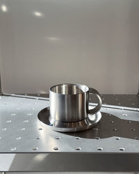 Stainless steel cup and saucer on a metallic surface with a neutral background